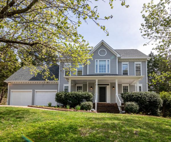 Two-story grey home with brick front steps, trimmed hedges on either side, green lawn, and a mature tree shading the front yard.